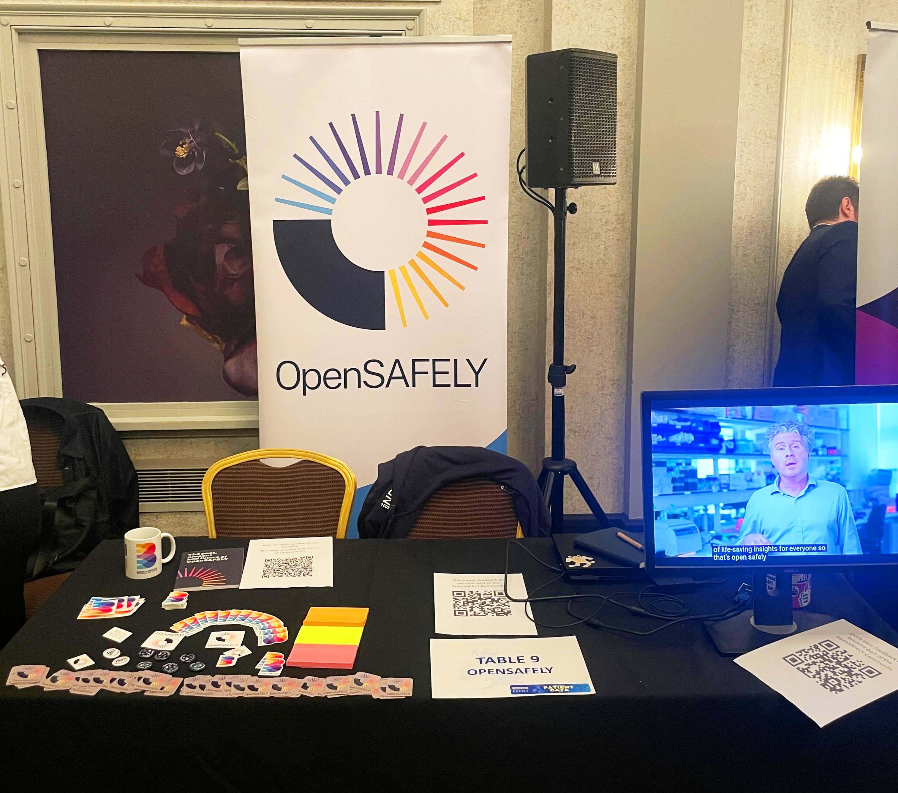 Our booth, featuring assorted Bennett and OpenSAFELY swag as well as a monitor showing the ‘robot video’ on loop.