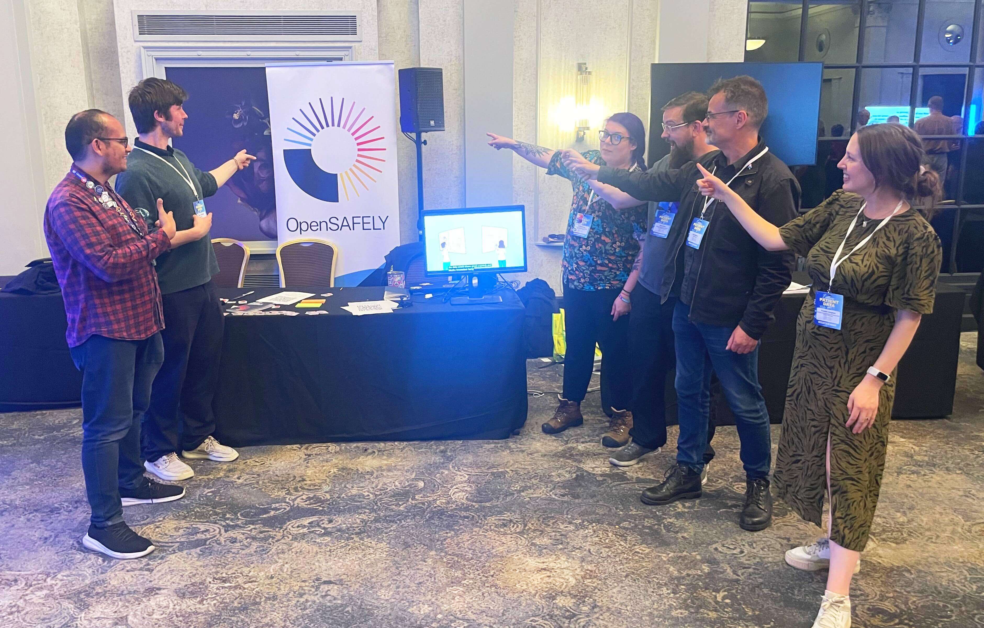 From left to right: Arif, John, Eli, Simon, Andy and Frederica all pointing at the OpenSAFELY banner behind our booth.
